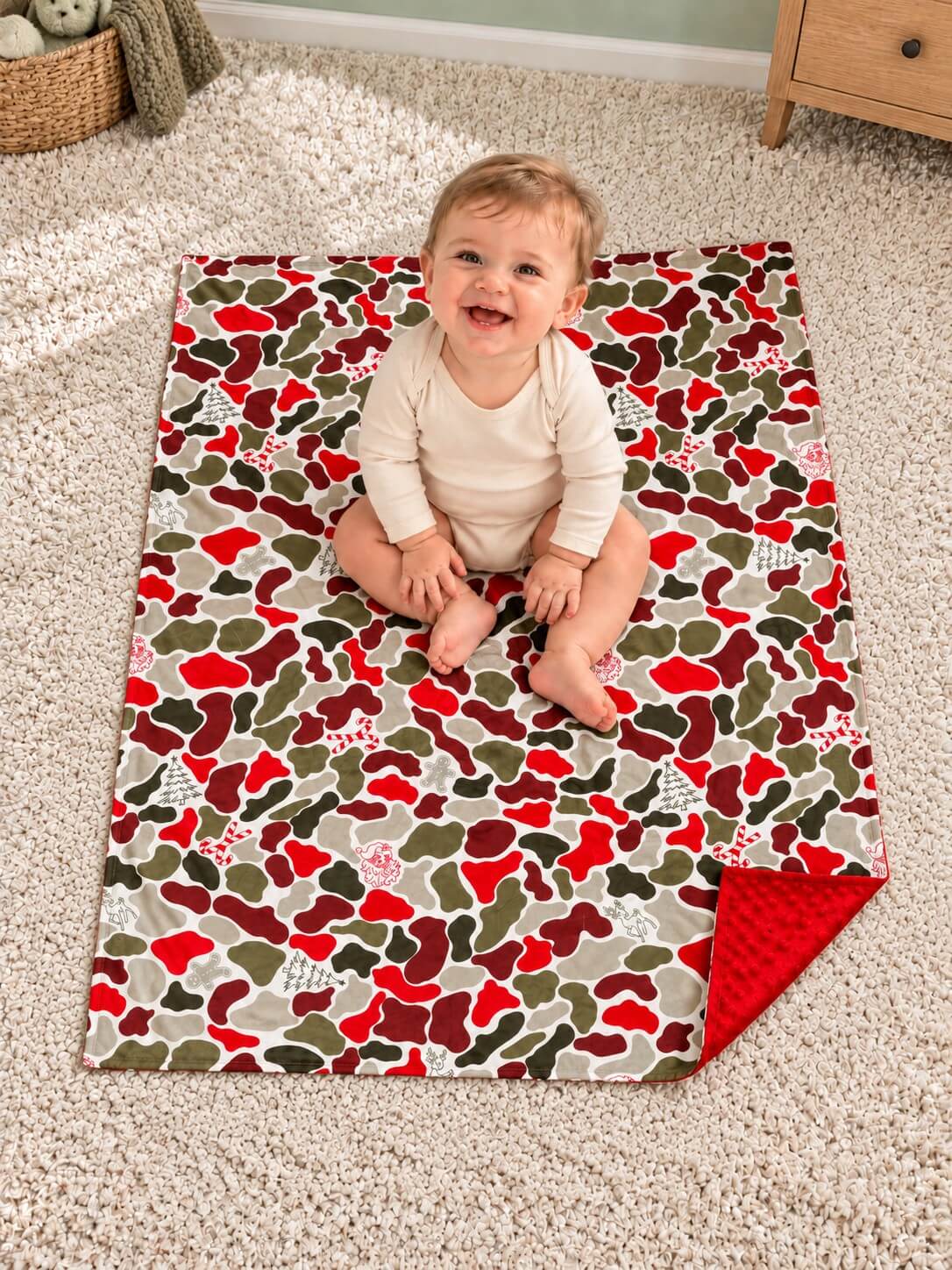 Christmas camouflage baby blanket with red minky dot backing spread on the floor beneath a baby, viewed from above