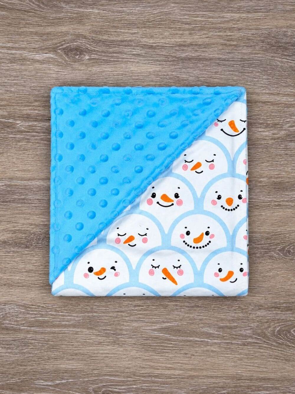 Blue snowman baby blanket with blue minky dot backing spread out beneath a seated baby